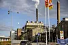 Billedet viser Sukkerfabrikken i Nakskov med fabrikken og en skorsten i baggrunden og to flag i forgrunden fra en nærliggende tankstation.
