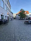 Billedet viser en del af Torvet, der er belagt med chaussesten. Man ser parkerede cykler på venstre side og en hvid bygning. I baggrunden ses flere bygninger, der huser forretninger.